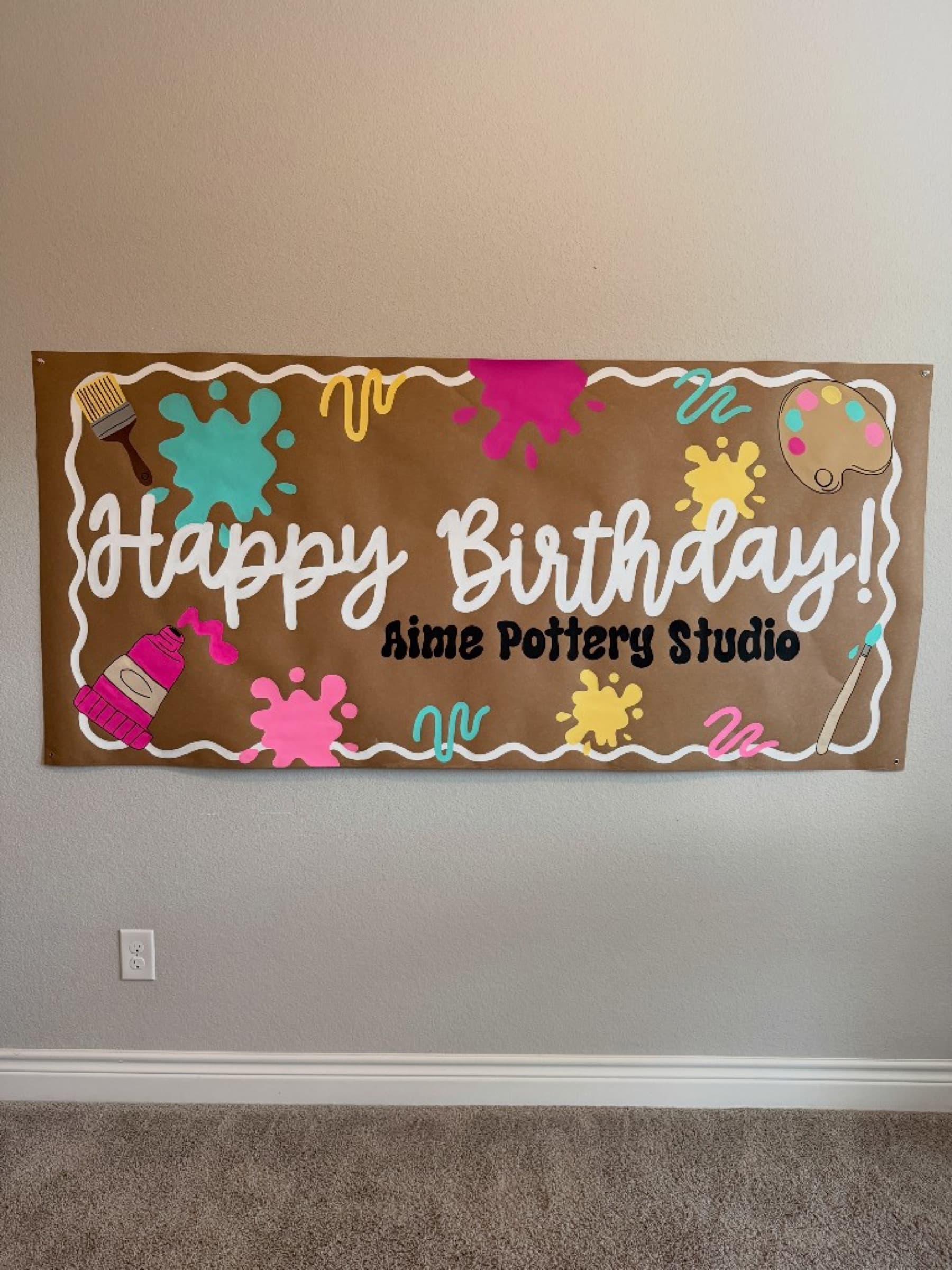 Kraft paper banner reading Happy Birthday Aime Pottery Studio with paint splats, a palette, brushes, and a paint tube