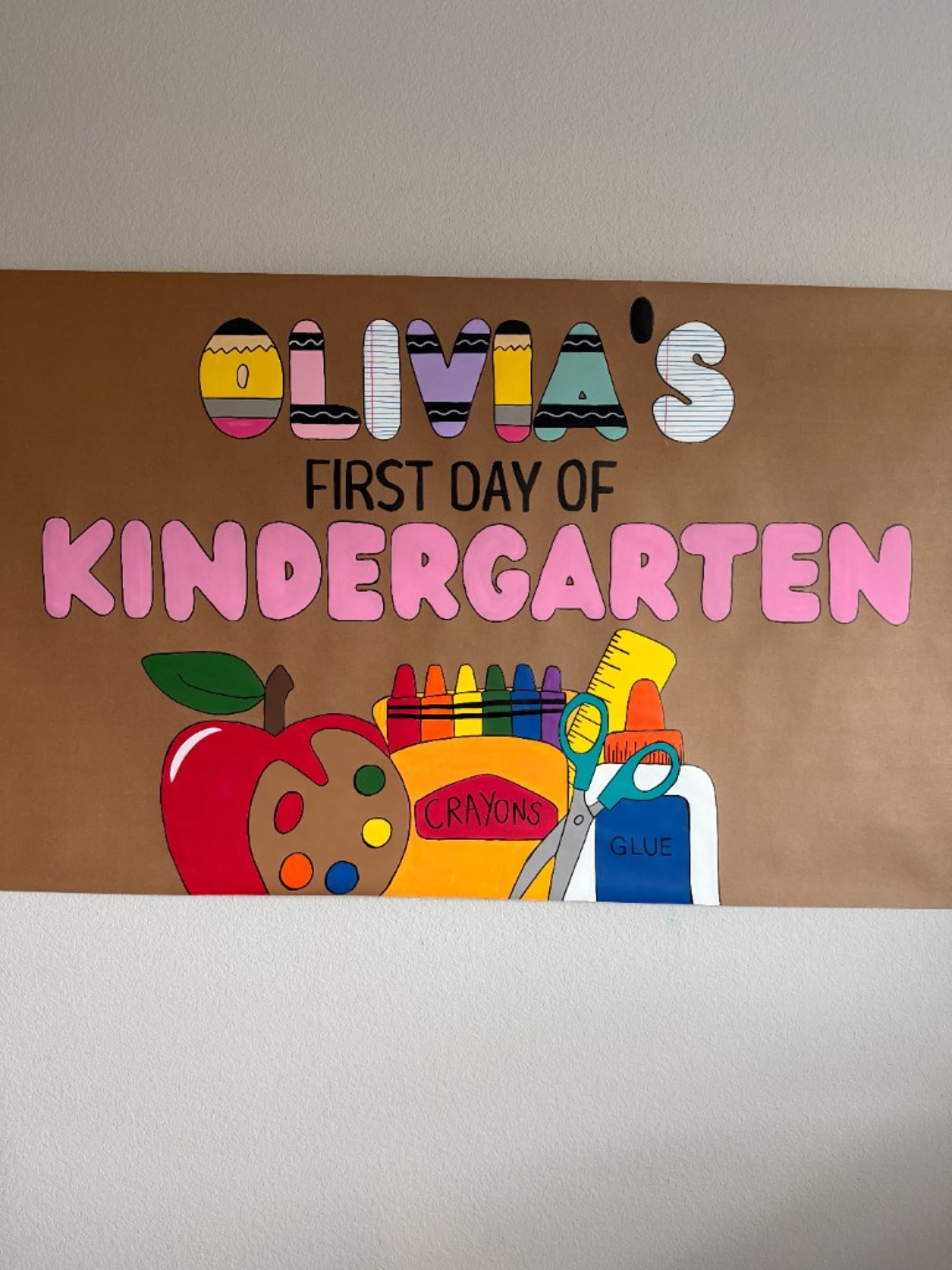 Kraft paper banner reading Olivia's First Day of Kindergarten with patterned letters, crayons, scissors, glue, and an apple