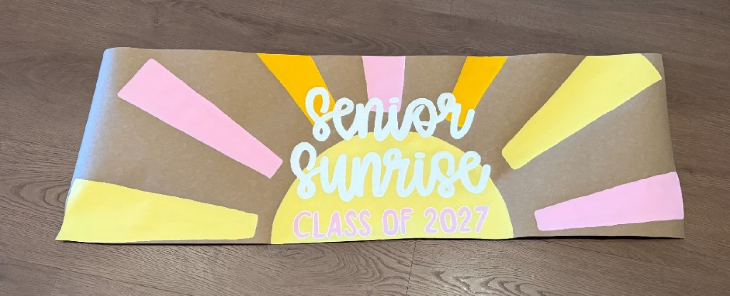 Wide Kraft paper banner reading Senior Sunrise Class of 2027 with a yellow sun and pink and yellow rays